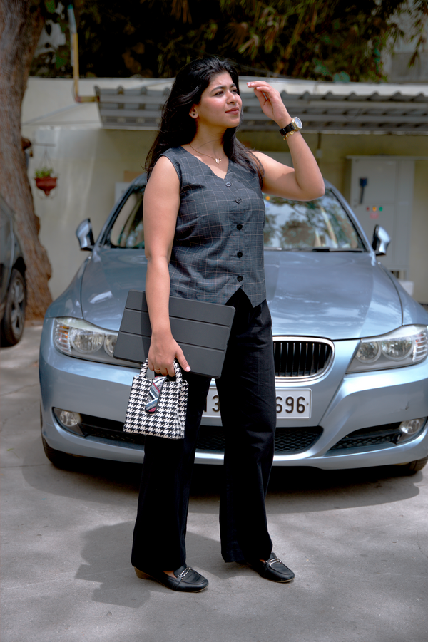 Woman wearing smart casual work wear black 9 to 9 pant and grandpa checks vest standing in front of a silver BMW car holding a checkered handbag.