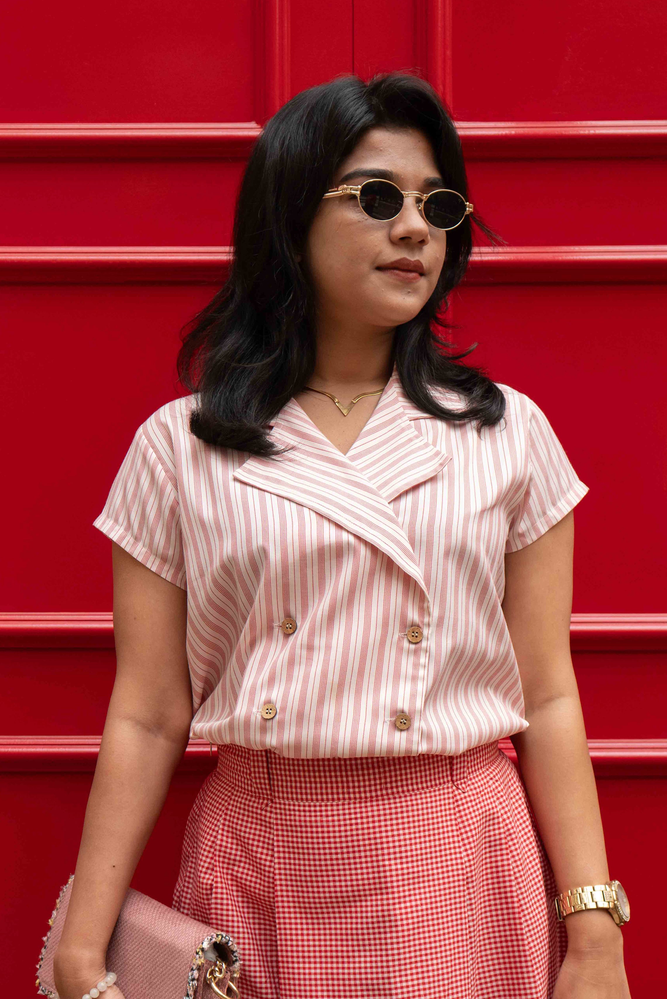 Woman wearing a smart casual work wear  double breasted blush stripe shirt and red pin check skirt against a red background