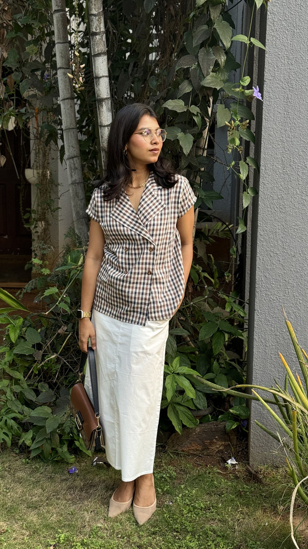 Woman wearing smart casual work wear cocoa checks double breasted shirt and white lady diana skirt standing outdoors with plants and a building in the background