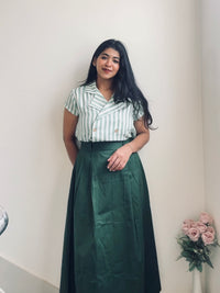 Front view of a woman wearing a smart casual work wear Match striped double breasted shirt and ever green skirt standing against a white wall with a vase of pink roses on a table.